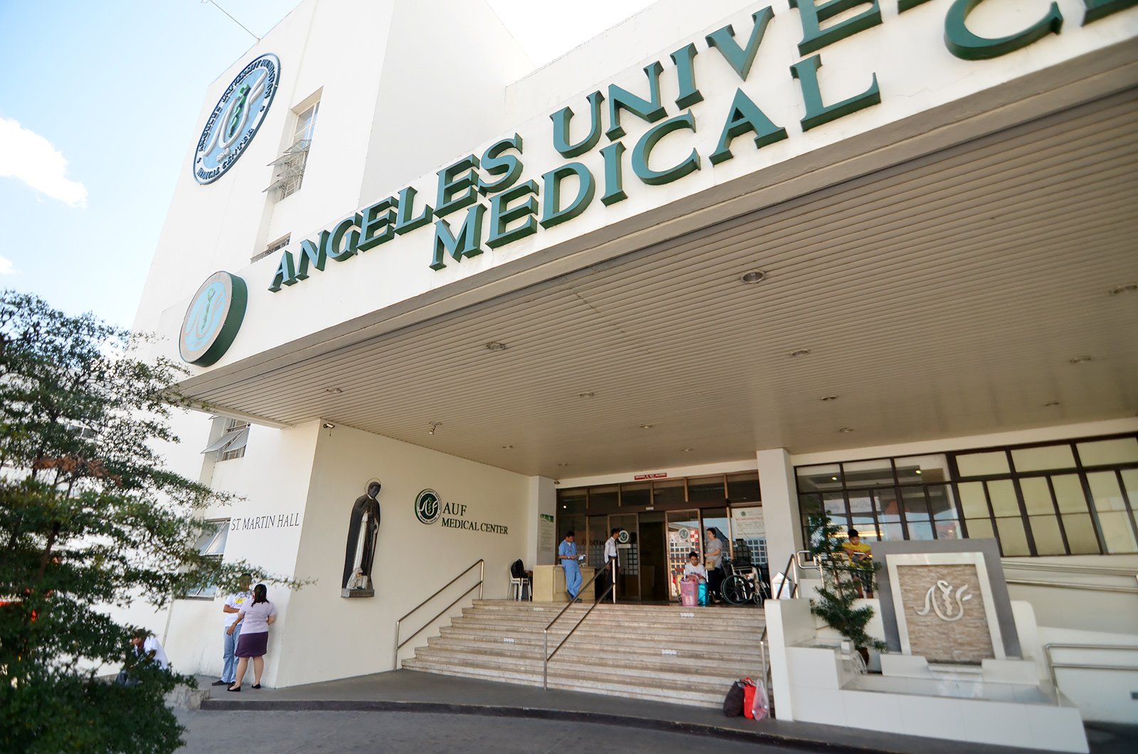 Angeles University Foundation Medical Center の正面入口。緑色の校章と施設名の看板が掲げられ、スタッフと来院者が出入りしている